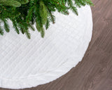 White quilted Christmas tree skirt with green tree branches on a wooden floor.