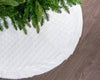 White quilted Christmas tree skirt with green tree branches on a wooden floor.