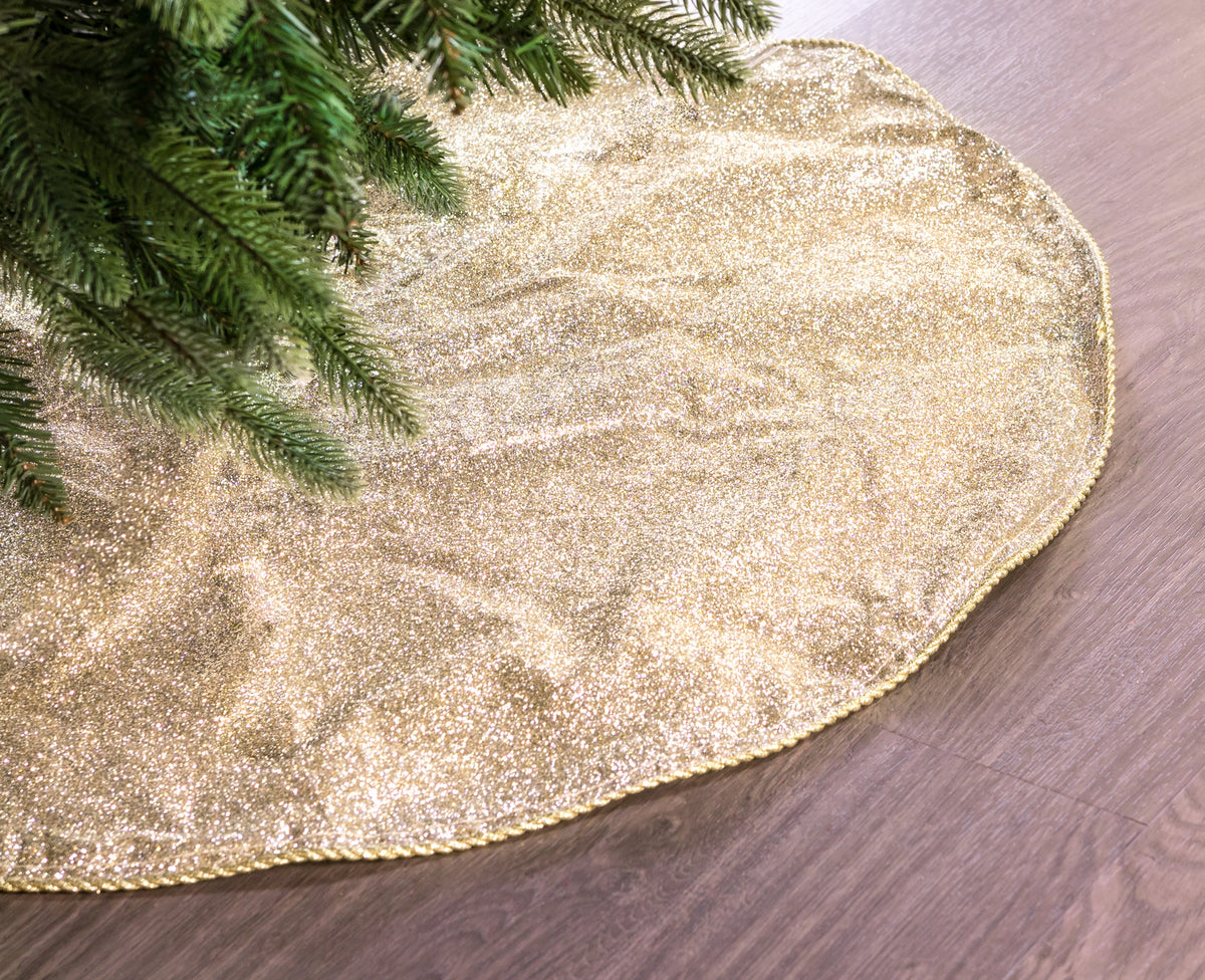 Gold glittery Christmas tree skirt on a wooden floor with a tree branch in the corner.
