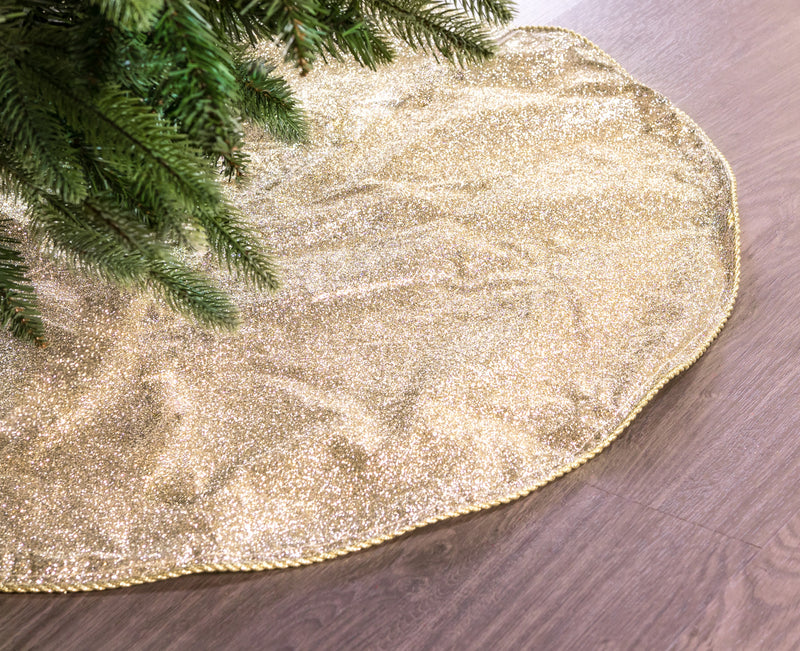 Gold glittery Christmas tree skirt on a wooden floor with a tree branch in the corner.