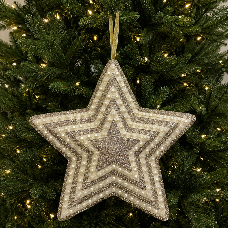 A large, five-pointed star Christmas ornament with concentric geometric patterns in beige and gray, hanging from a gold ribbon on a lit green Christmas tree.