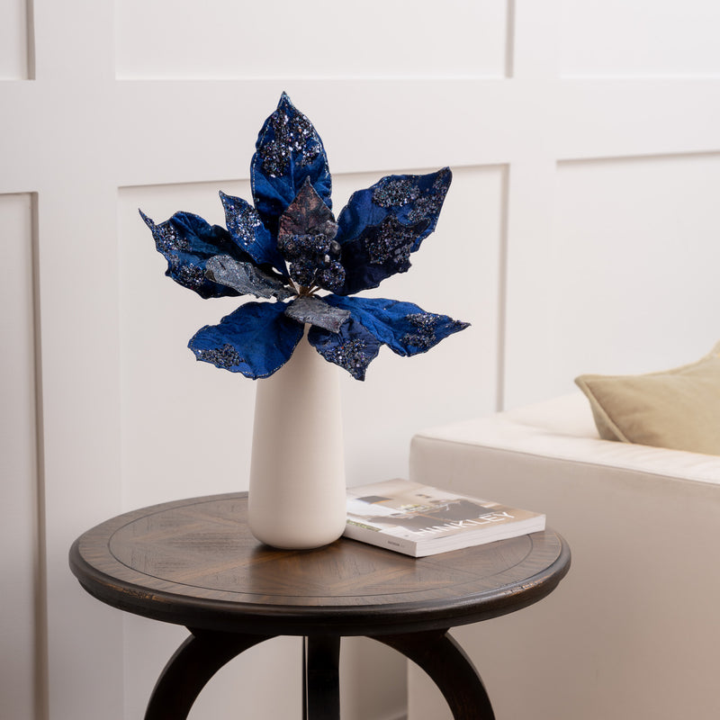 Decorative blue flower arrangement in a white vase on a wooden side table with a neutral background.