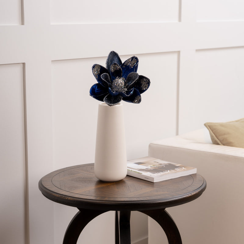 Decorative vase with a blue flower on a wooden side table against a white wall.