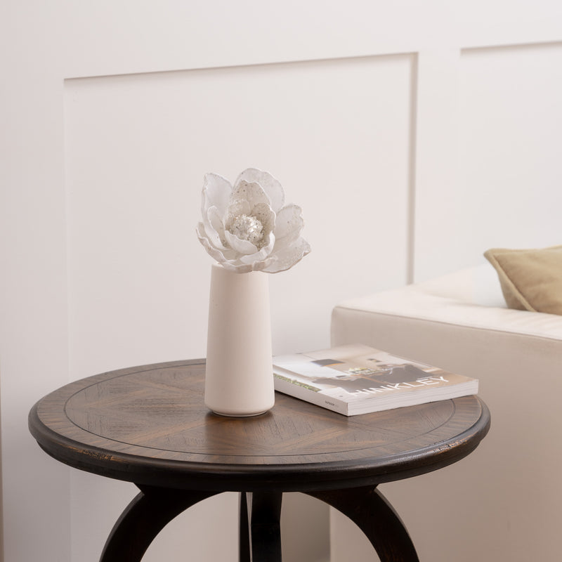 White vase with white glitter flower and book against a neutral wall.
