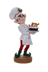 Elf figurine in a chef's outfit holding a cake with 'Joy' on it, on a white background.