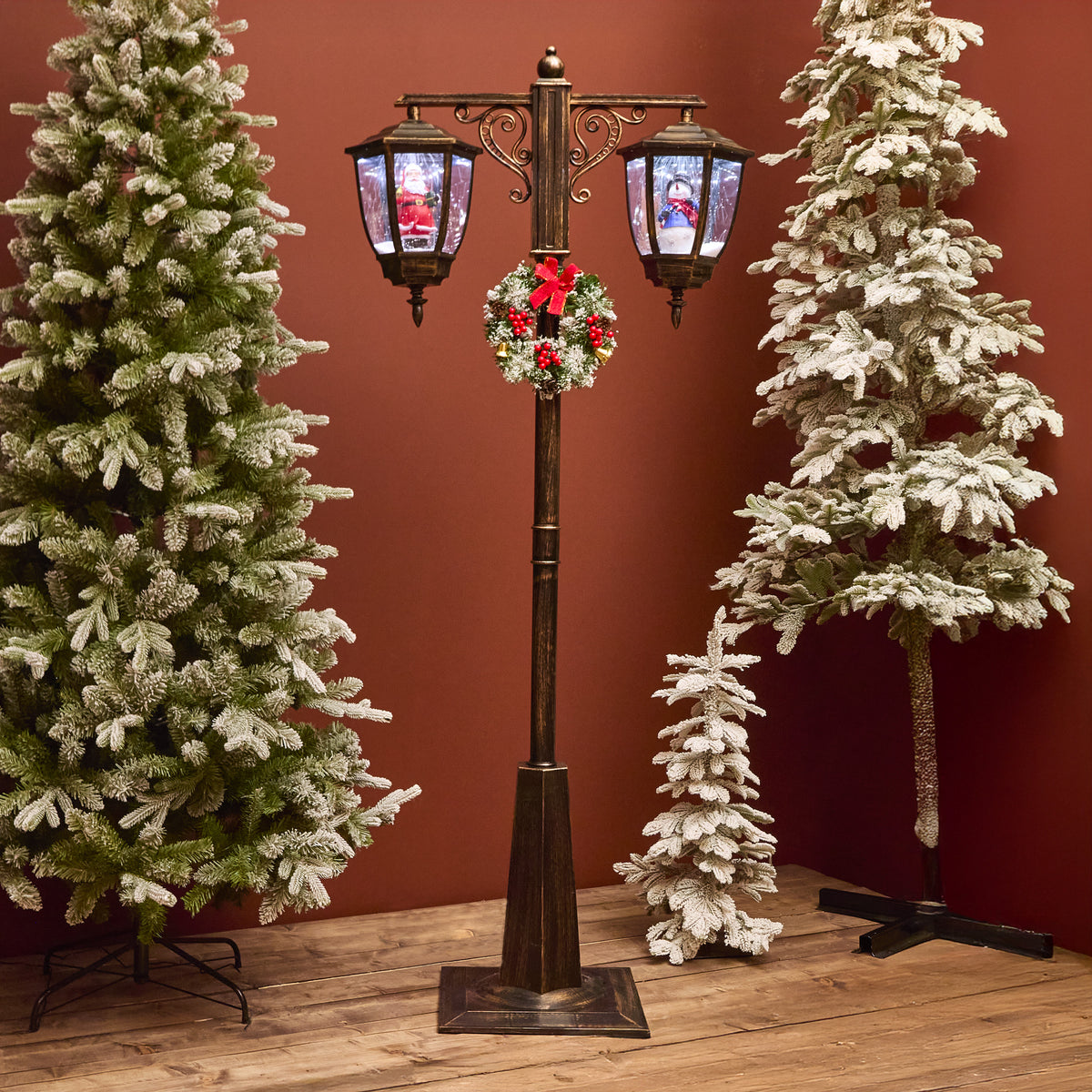 Decorative Christmas street lamp with wreath surrounded by snow-covered trees against a red wall.