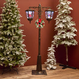 Decorative Christmas street lamp with wreath surrounded by snow-covered trees against a red wall.