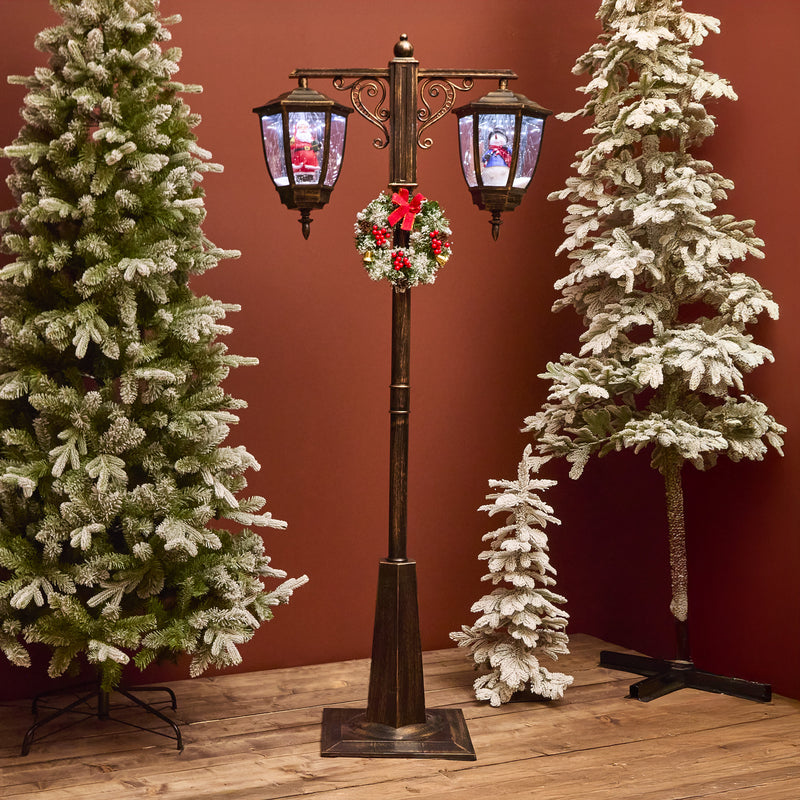 Decorative Christmas street lamp with wreath surrounded by snow-covered trees against a red wall.