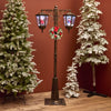 Decorative Christmas street lamp with wreath surrounded by snow-covered trees against a red wall.