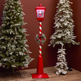 Decorative Christmas lamp post with candy cane design and wreath, surrounded by snow-dusted trees.