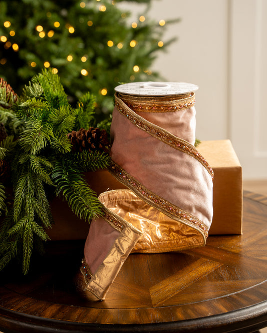 Pink ribbon with gold trim on a wooden table next to a Christmas tree.