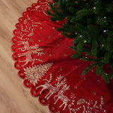 Red Christmas tree skirt with gold reindeer and floral patterns next to a decorated tree on a wooden floor.