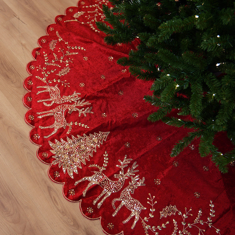 Red Christmas tree skirt with gold reindeer and floral patterns next to a decorated tree on a wooden floor.