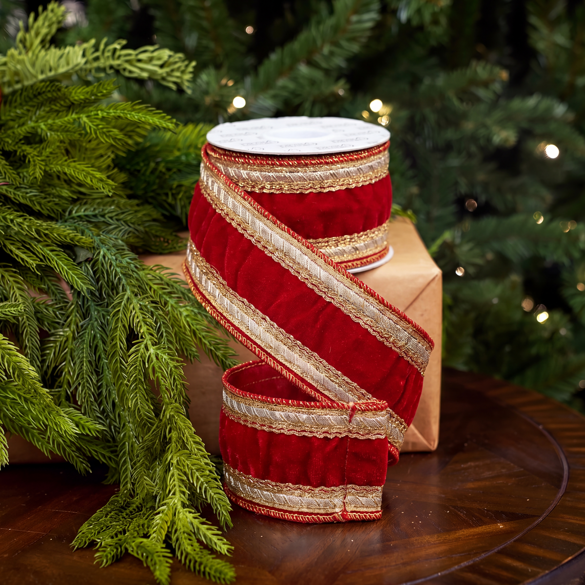 A roll of red velvet holiday ribbon with metallic gold stripes and trim, wrapped around a kraft gift box on a wooden table with pine branches and a blurred Christmas tree in the background.