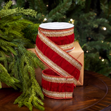 A roll of red velvet holiday ribbon with metallic gold stripes and trim, wrapped around a kraft gift box on a wooden table with pine branches and a blurred Christmas tree in the background.