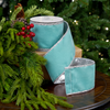 A roll of teal velvet ribbon with silver glitter trim, displayed on a wooden surface next to green pine branches and red berries, with a blurred Christmas tree in the background.