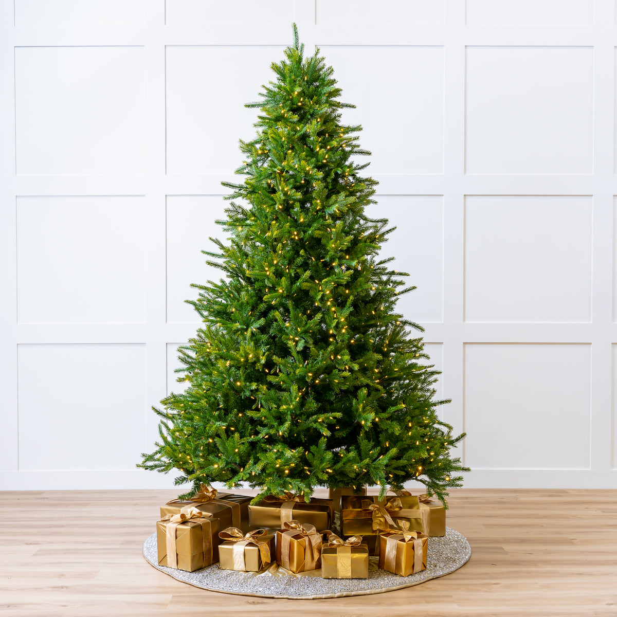 Decorated Christmas tree with gold presents on a wooden floor against a white paneled wall.