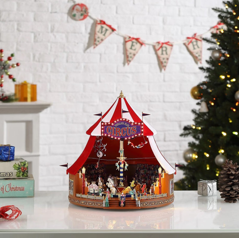 Decorative circus tent model on a table with Christmas tree and decorations in the background.