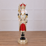 A detailed, painted nutcracker soldier figurine standing on an ornate round base. The figurine features a red and gold crown, a white beard, a silver and gold decorated coat, red trousers, black boots, and holds a gold spear.