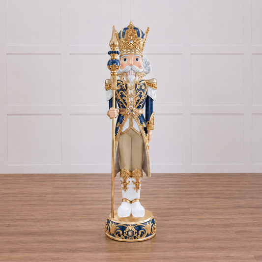 A decorative nutcracker figurine dressed as a royal prince, standing on a round ornate base and holding a spear. The figure has a white face and beard, a gold crown and trim, navy blue sleeves, and beige pants, photographed against a plain white background.